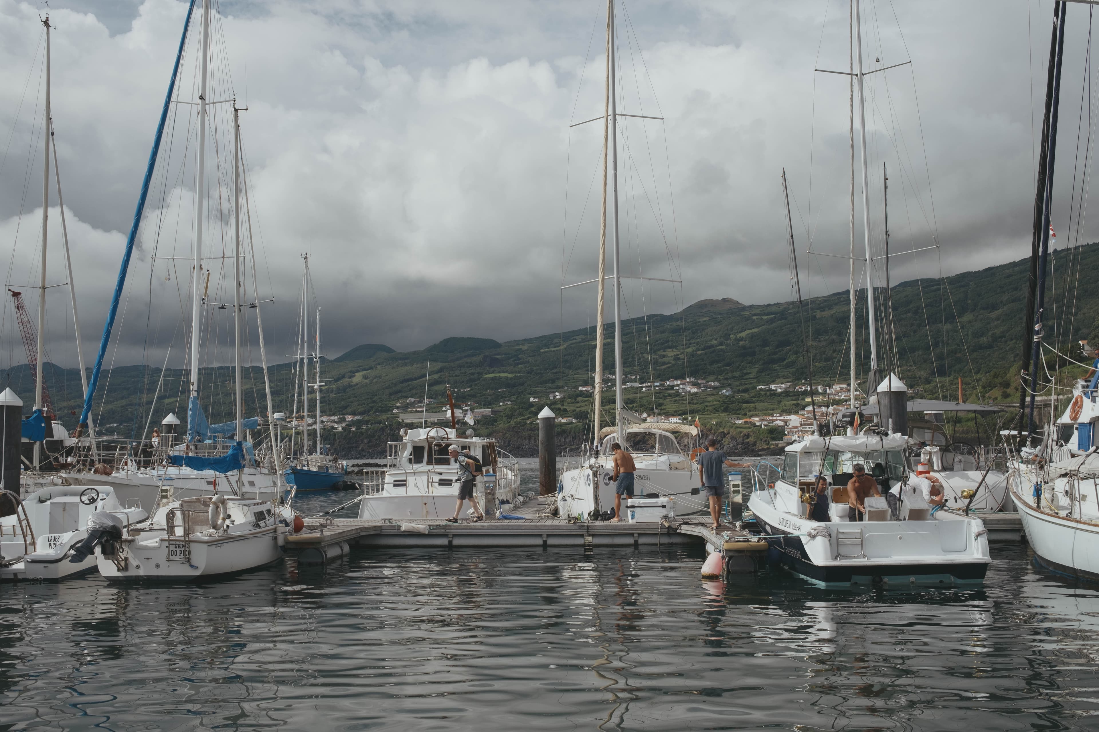 Marina in the Azores with white sailboats and yachts moored at wooden docks, their tall masts creating a forest of rigging against an overcast sky. People can be seen on several boats preparing for departure or maintenance. Green volcanic hills rise in the background with scattered white buildings of a coastal town nestled among the slopes. The calm harbor water reflects the boats and cloudy sky.