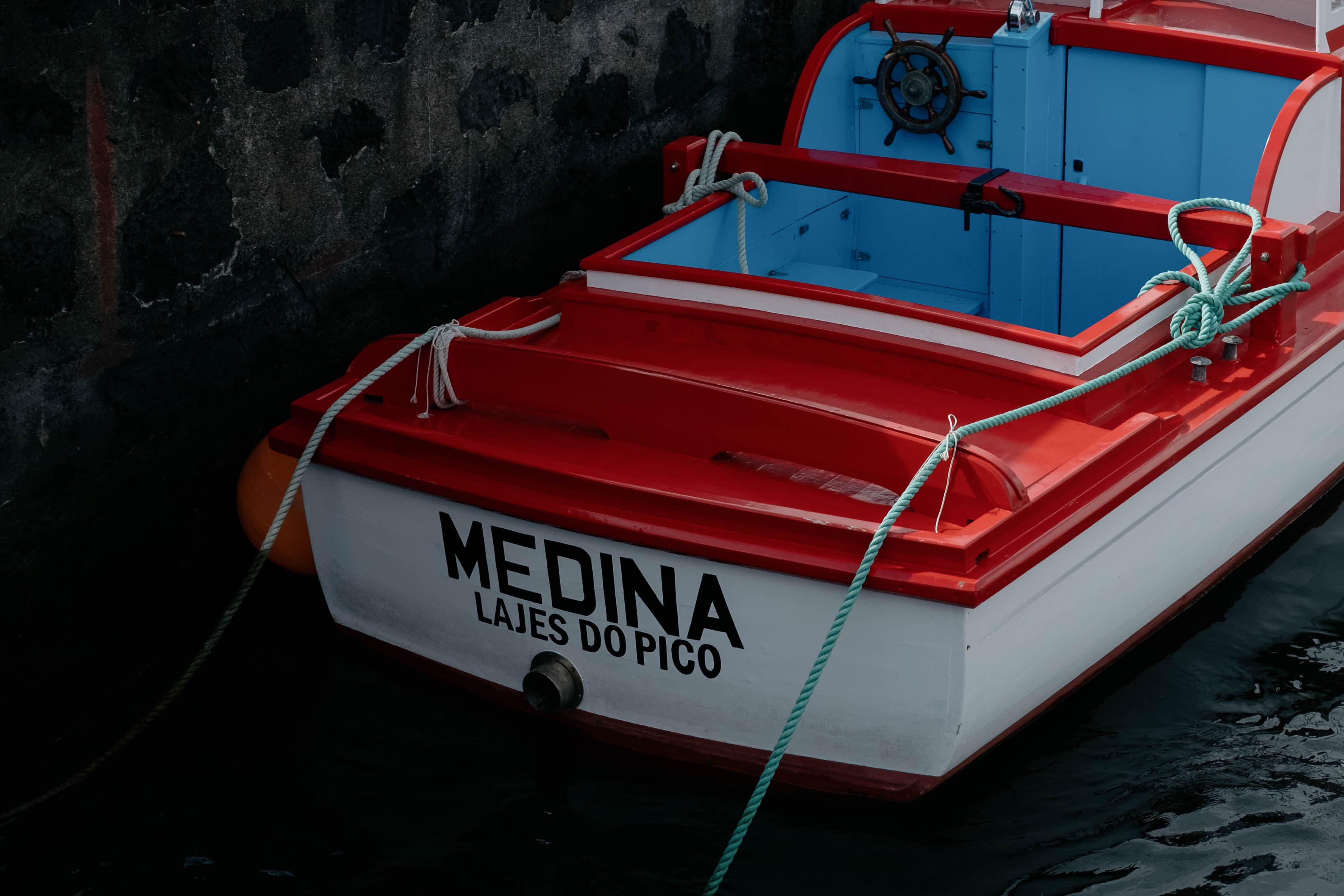 Traditional Azorean fishing boat named 'MEDINA LAJES DO PICO' with a white hull and bright red trim, moored with green rope against dark volcanic rocks. The boat features a blue interior deck area with a black ship's wheel, coiled ropes, and classic fishing boat hardware. The contrast between the colorful vessel and the black lava rock shoreline is striking.