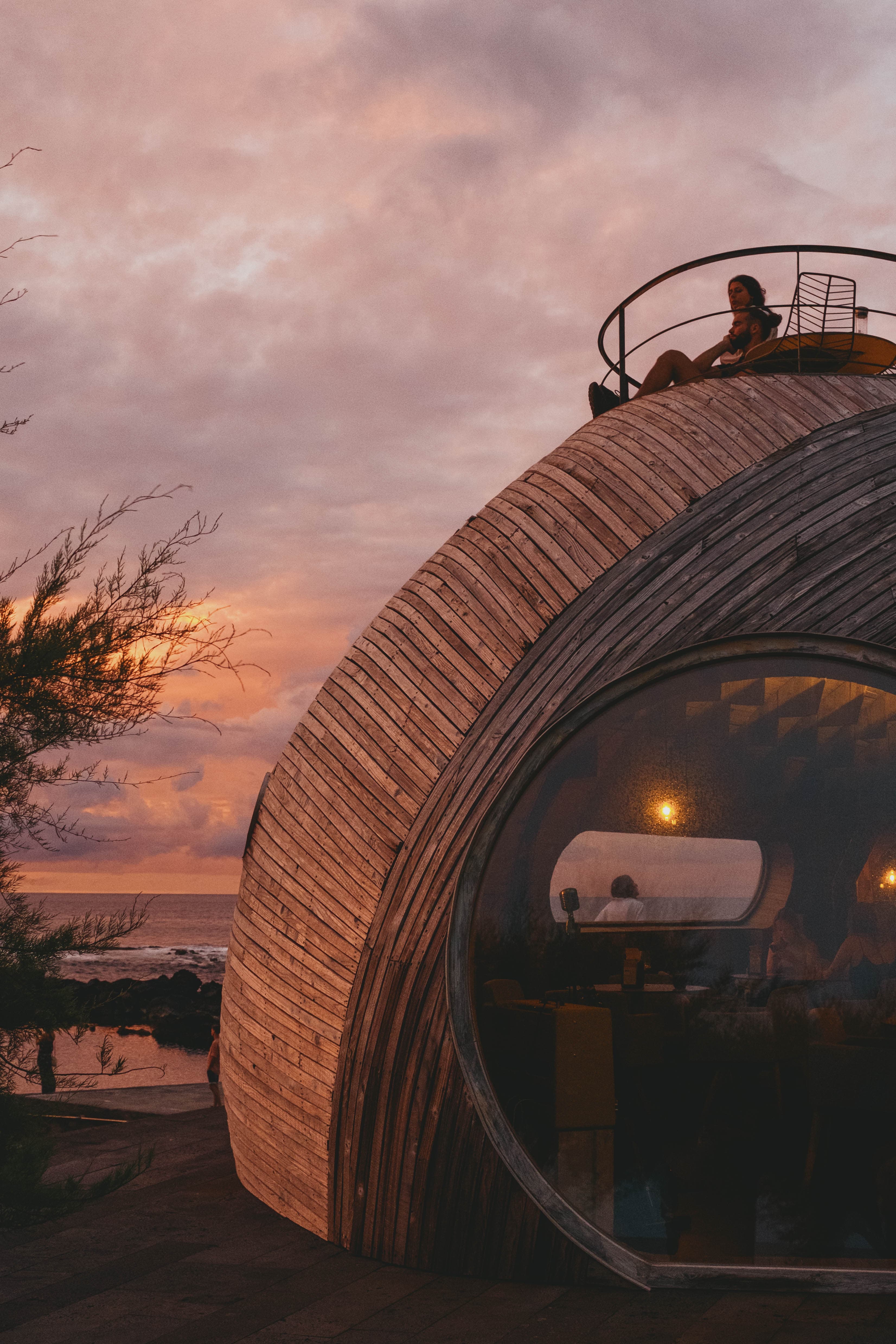 Curved wooden architecture of Cella Bar with a person relaxing on the rooftop terrace during golden hour sunset, overlooking the Atlantic Ocean with warm pink and orange skies