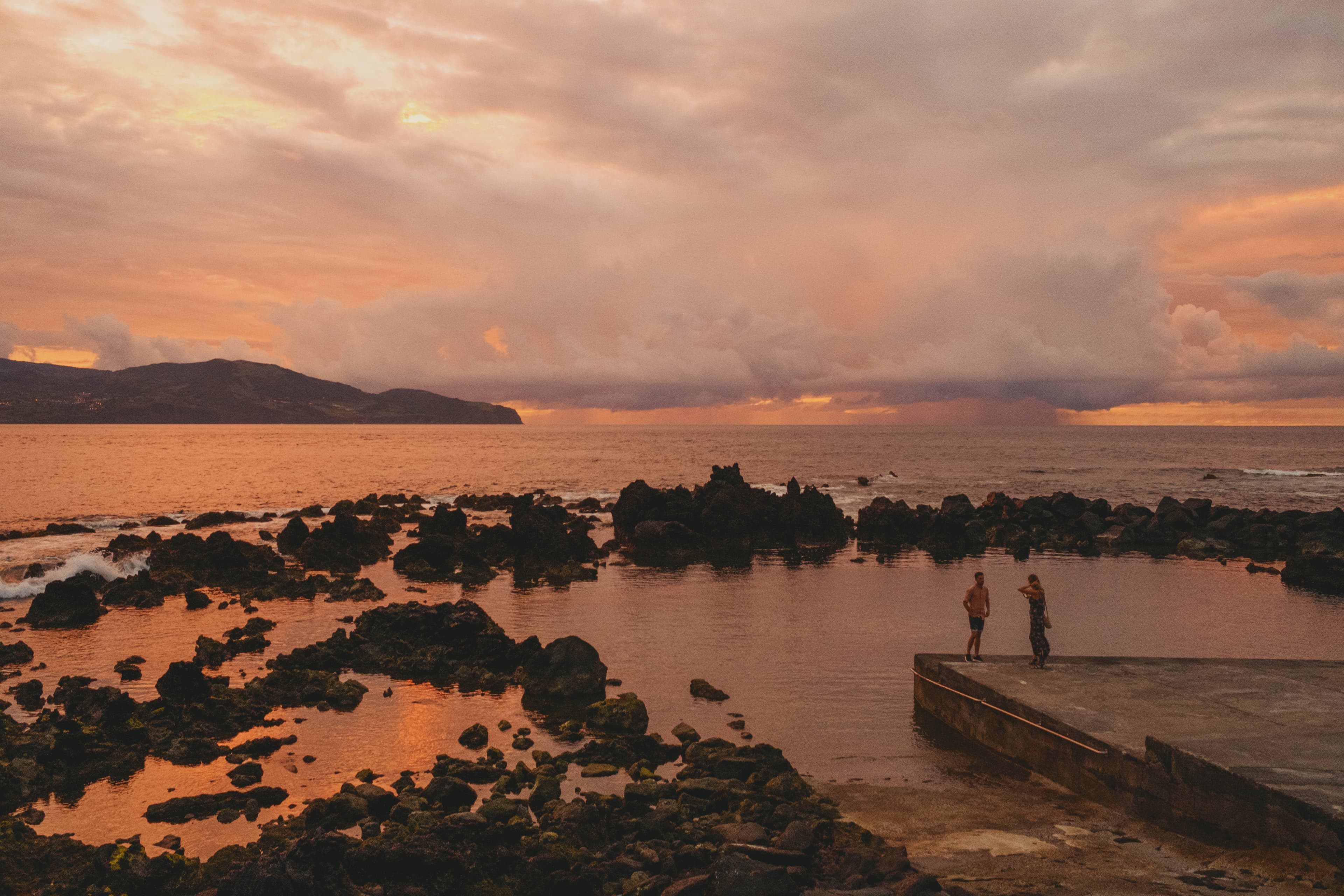Two people standing on a concrete pier at Cella Bar, surrounded by dramatic volcanic rocks and tide pools reflecting golden sunset light, with mountains silhouetted across the ocean
