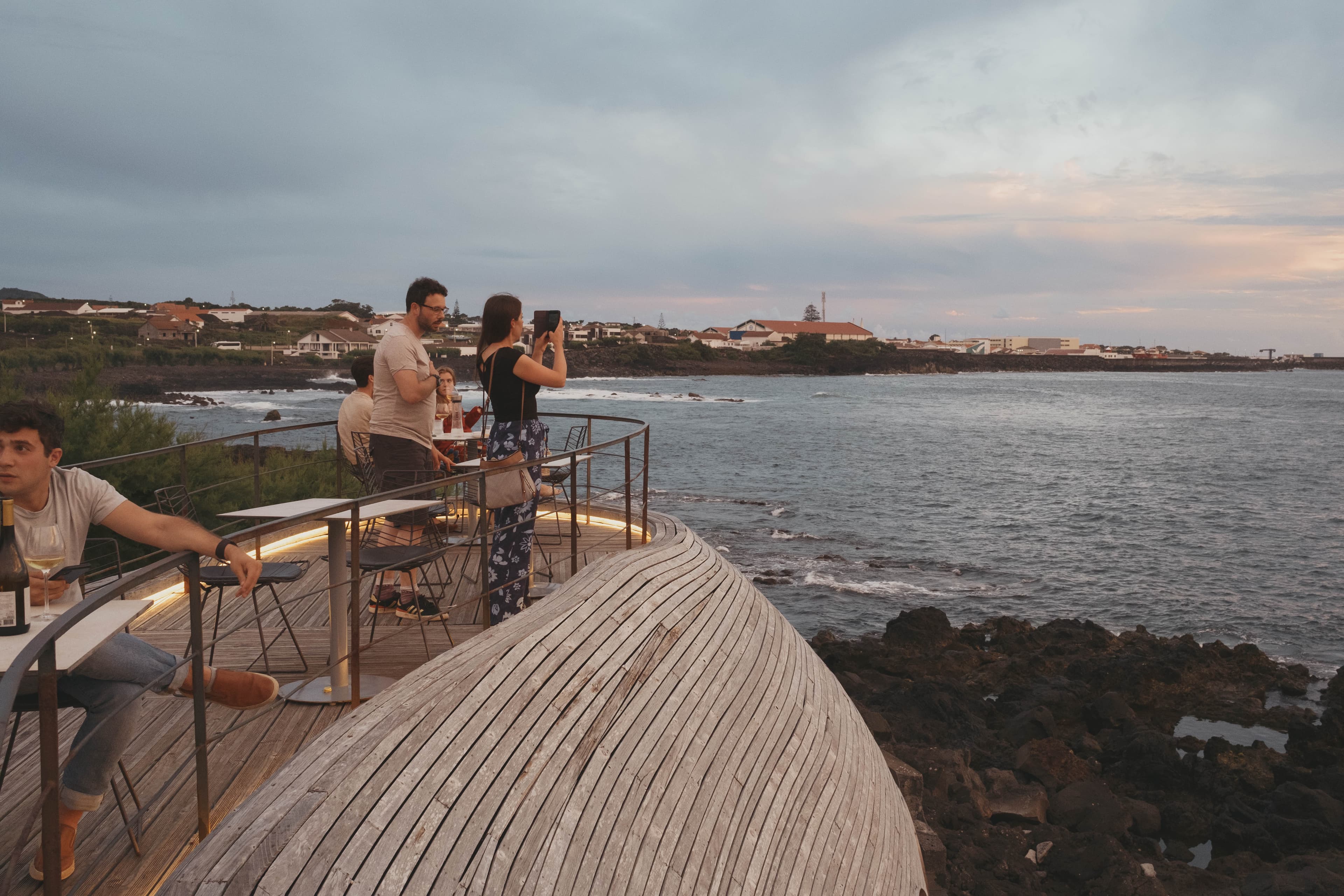 Guests enjoying drinks on the curved wooden deck at Cella Bar with ocean views of the volcanic coastline and traditional Portuguese village buildings in the background during evening hours