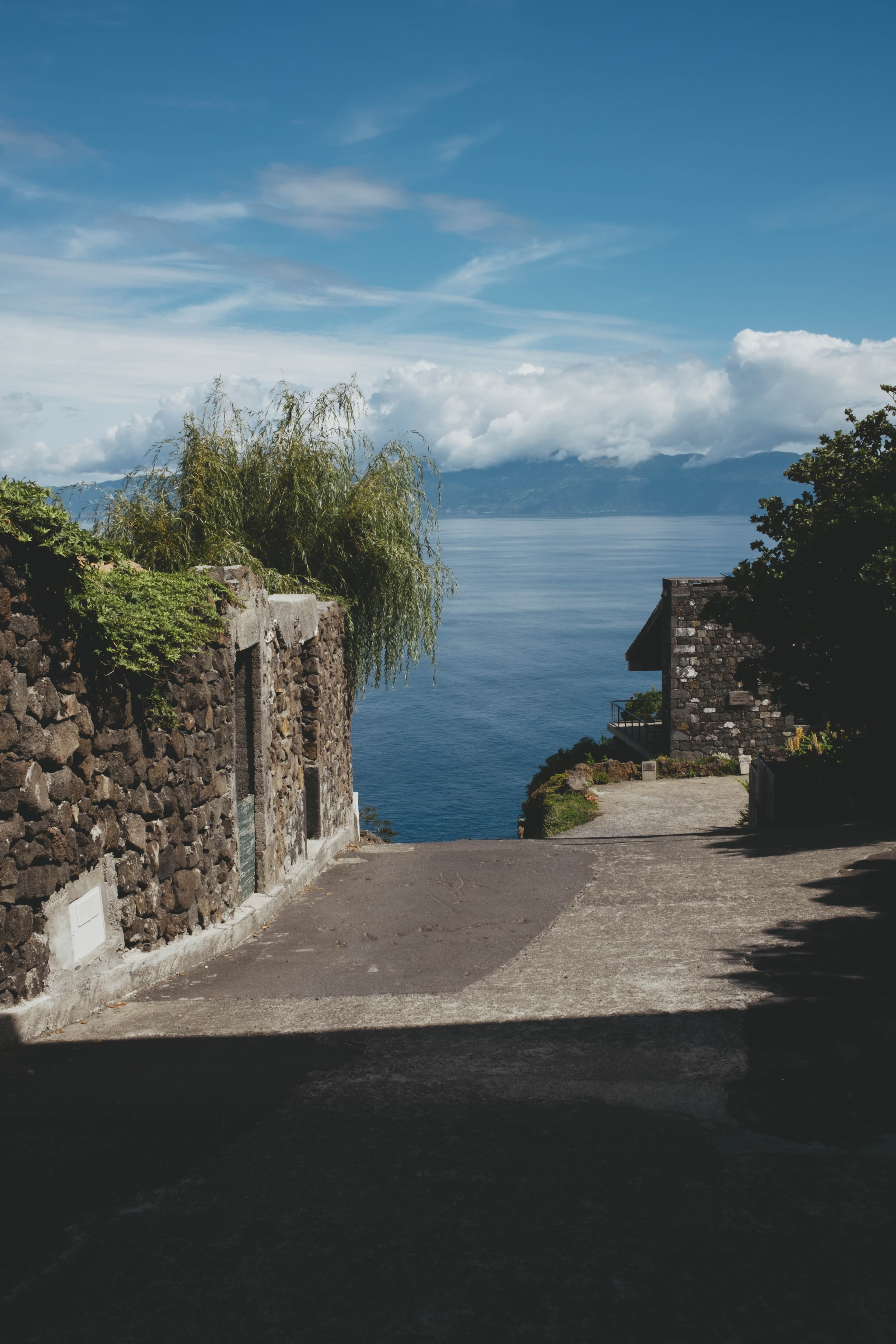 Coastal view in the Azores showing traditional stone buildings with volcanic rock walls flanking a concrete pathway that leads toward the Atlantic Ocean. A weeping willow tree grows between the structures, with lush green vegetation surrounding the buildings. The deep blue ocean stretches to distant mountainous islands partially obscured by white clouds under a bright blue sky.