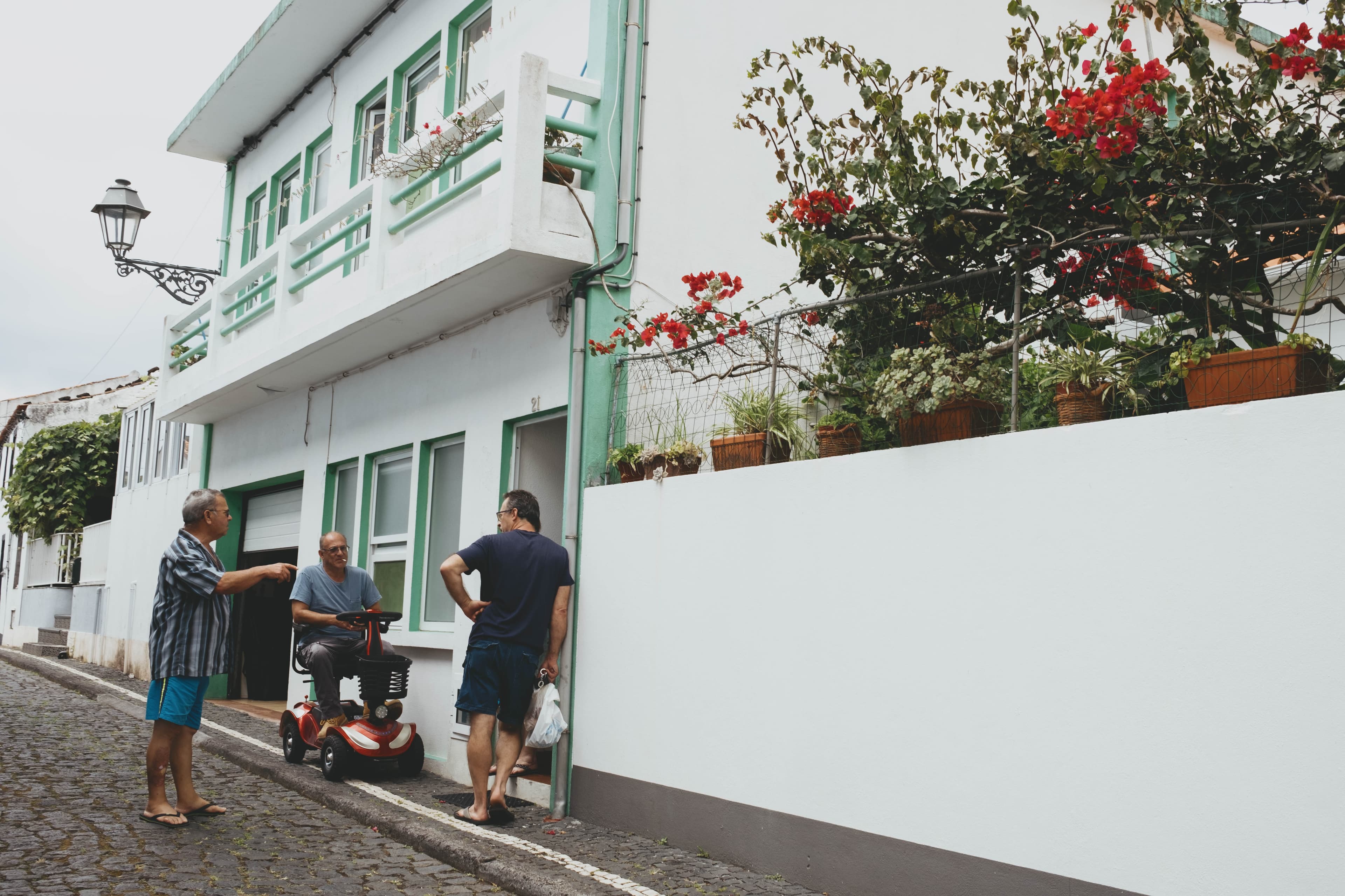 Three elderly men socializing on a cobblestone street in the Azores outside a traditional white house with mint green window shutters and trim. One man sits on a red mobility scooter while the other two stand chatting. The two-story home features a decorative iron street lamp and is adorned with vibrant red bougainvillea cascading over a white garden wall. Wooden planters and lush greenery complete the charming Portuguese island streetscape.