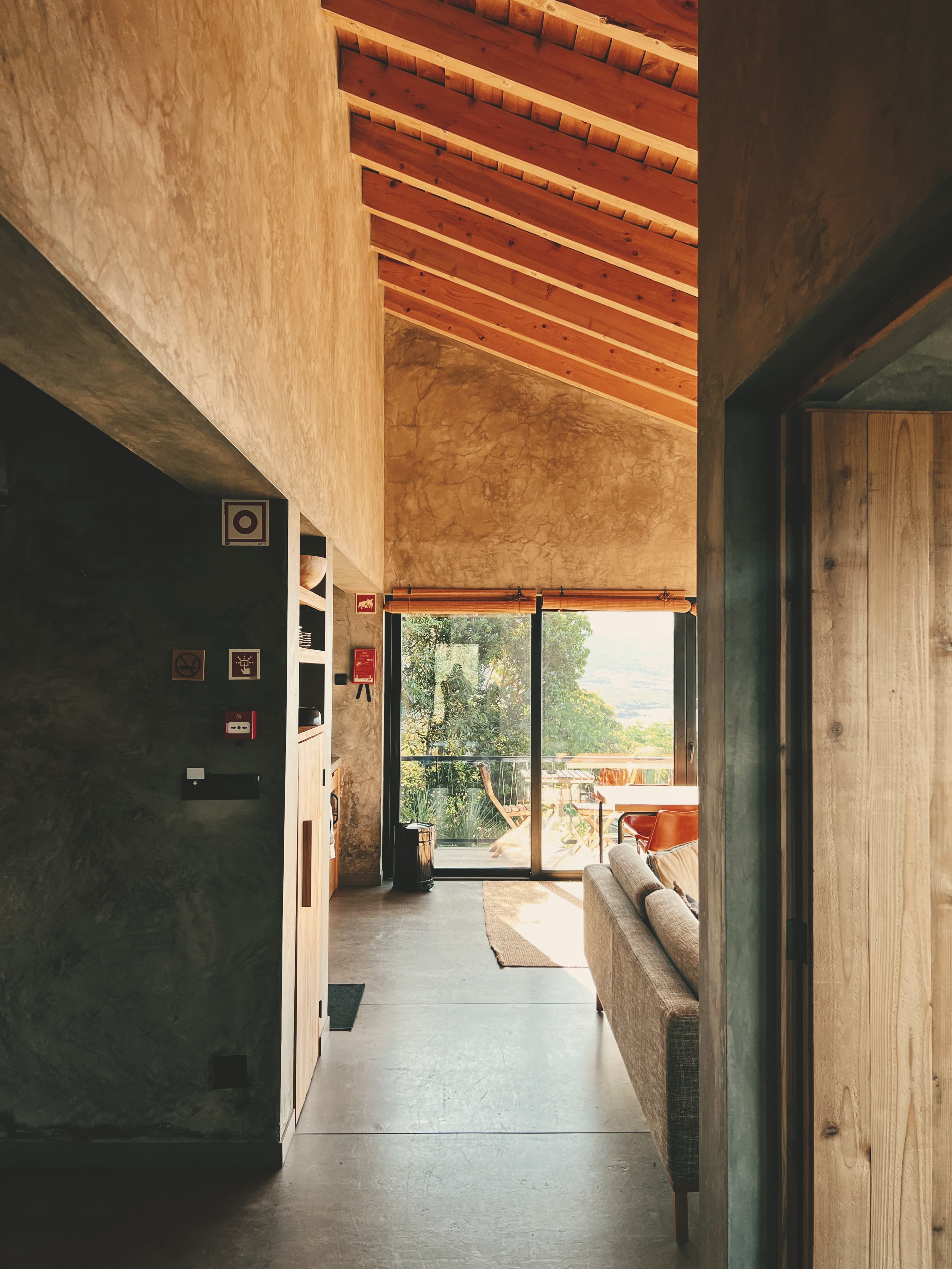 Modern living space with exposed wooden ceiling beams, concrete floors, and floor-to-ceiling windows opening to a terrace with mountain and ocean views