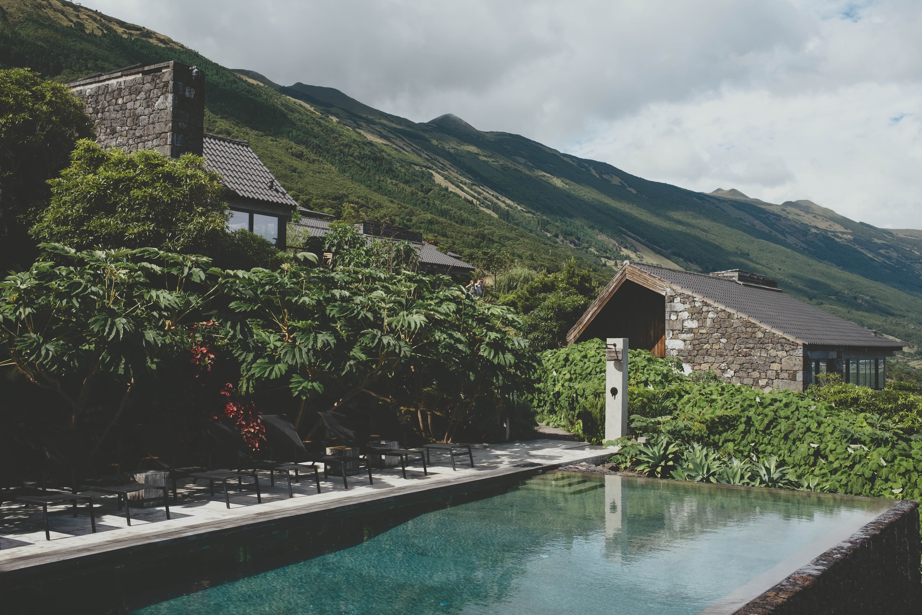 Modern stone and wood houses with terracotta roofs nestled against green mountain slopes, featuring a turquoise swimming pool and landscaping under cloudy skies