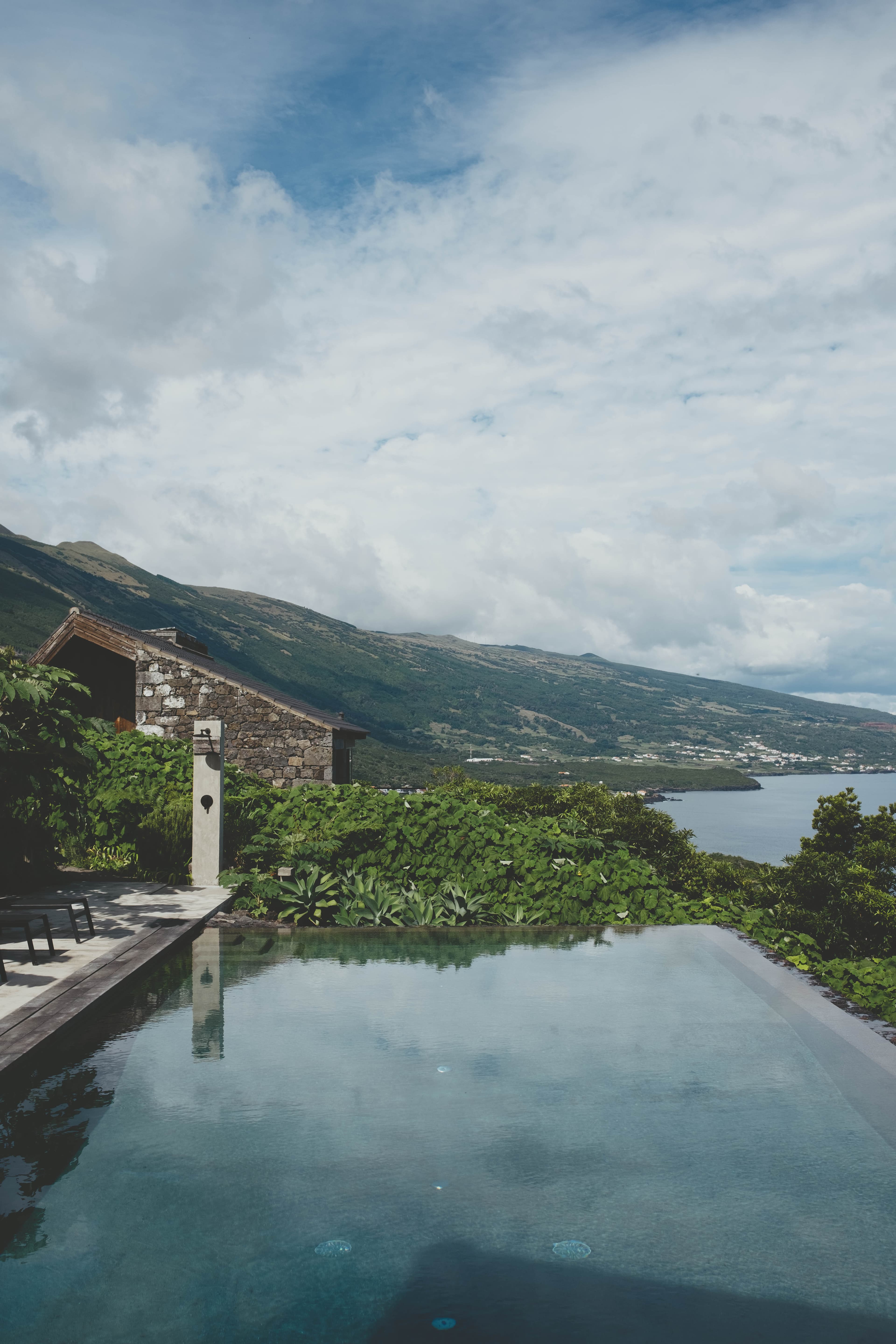 Infinity pool reflecting cloudy skies overlooks a coastal bay, with stone villa and lush gardens set against green mountain slopes