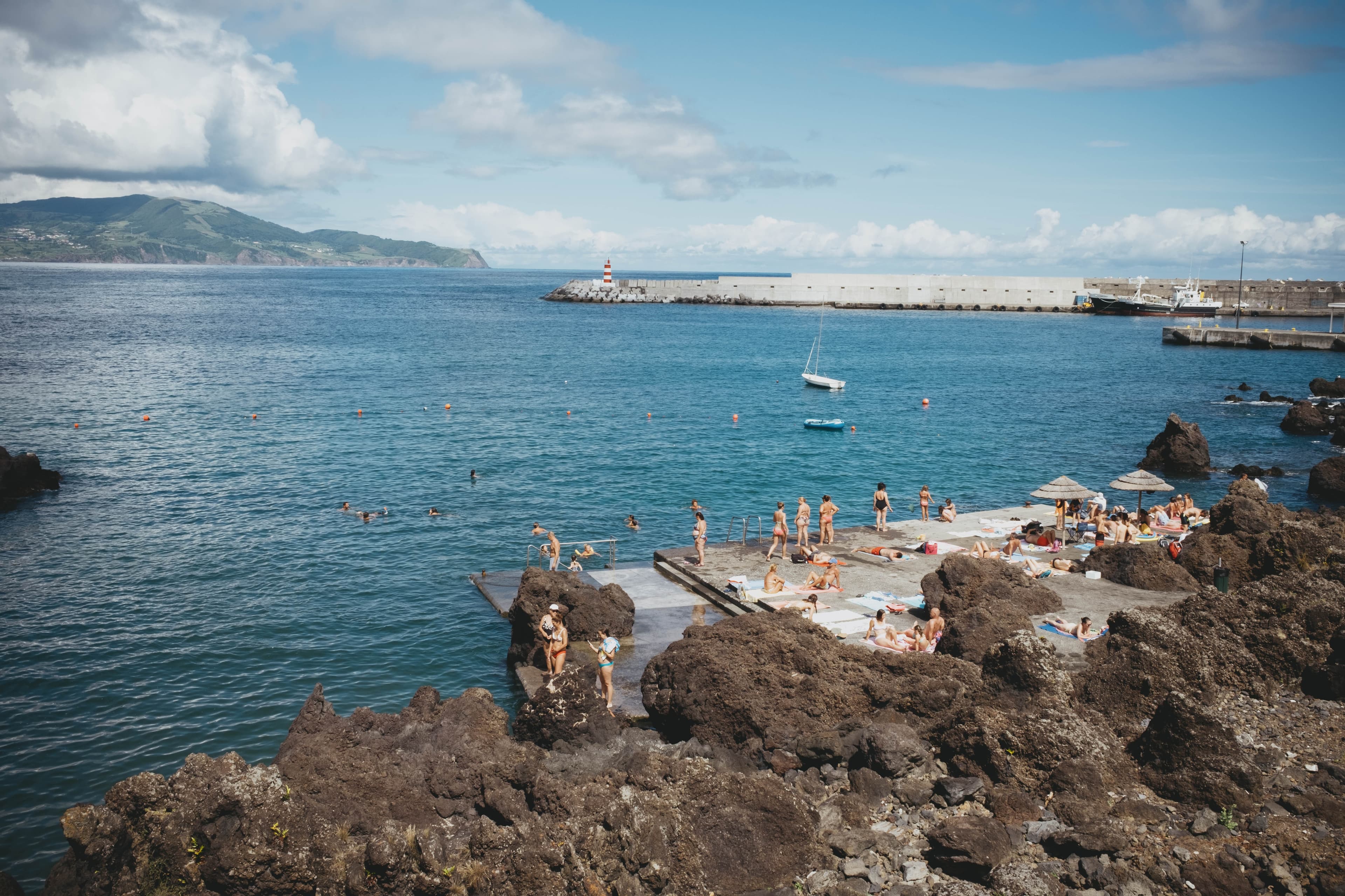 Swimmers and sunbathers at Madalena's natural lava rock pools on Pico Island, Azores, with the town's red and white striped lighthouse on the harbor breakwater and green mountains across the water
