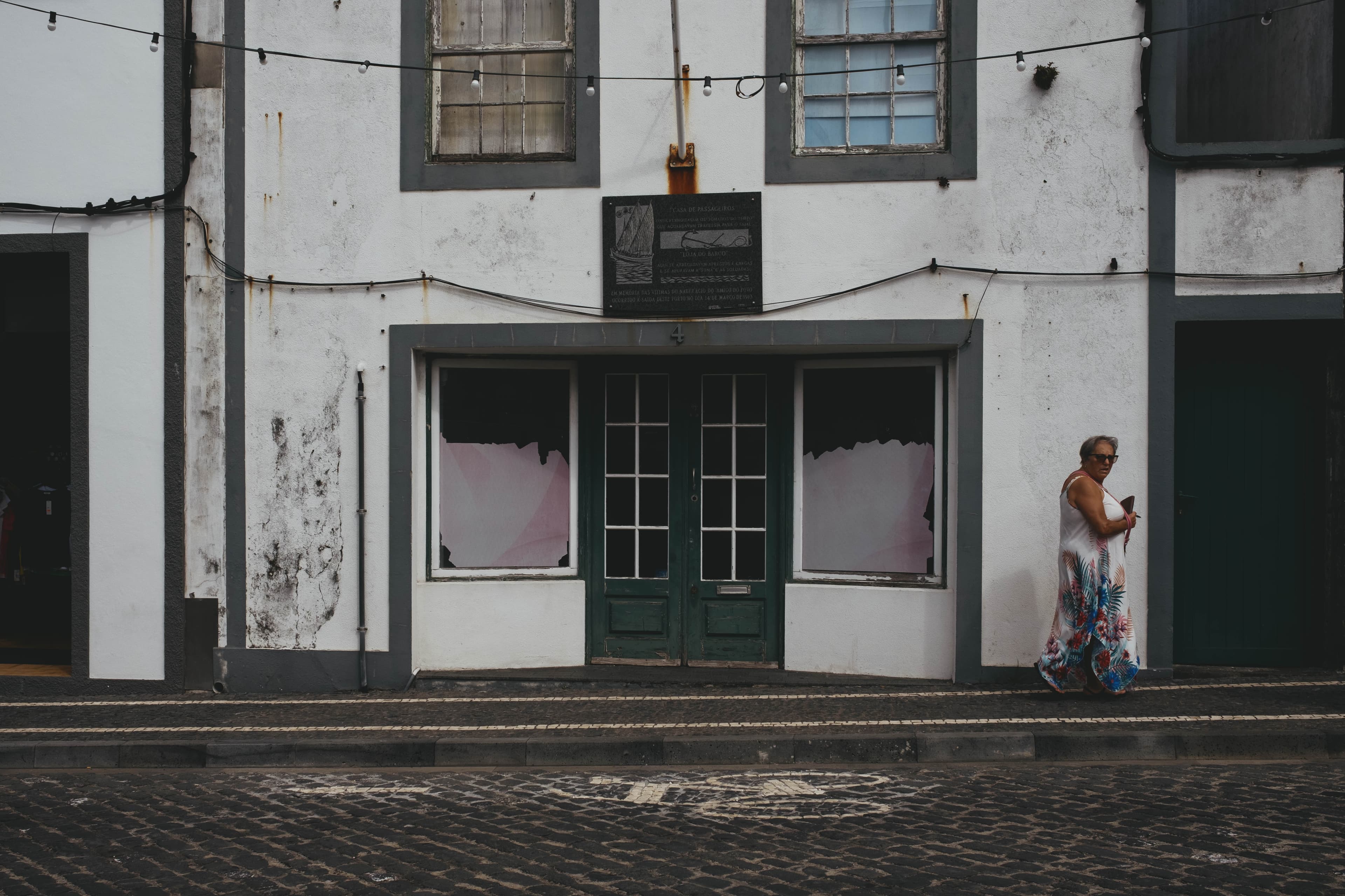 Woman in a floral dress walking past traditional white-washed buildings with dark green trim on a cobblestone street with string lights overhead and weathered storefronts