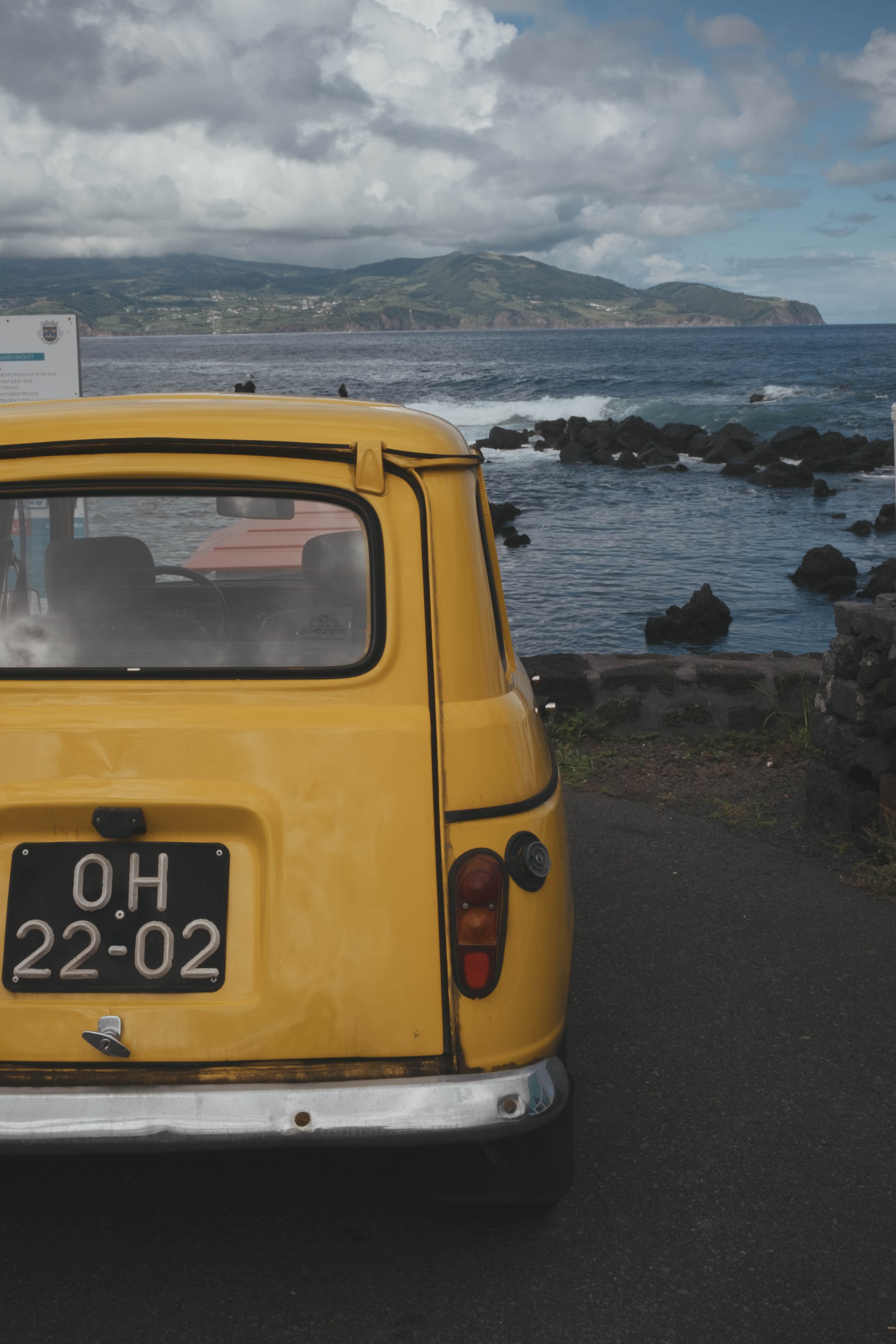 Yellow vintage Volkswagen van with Portuguese license plate parked overlooking the Atlantic Ocean on Pico Island, Azores, with volcanic rock coastline and green mountains under dramatic cloudy skies