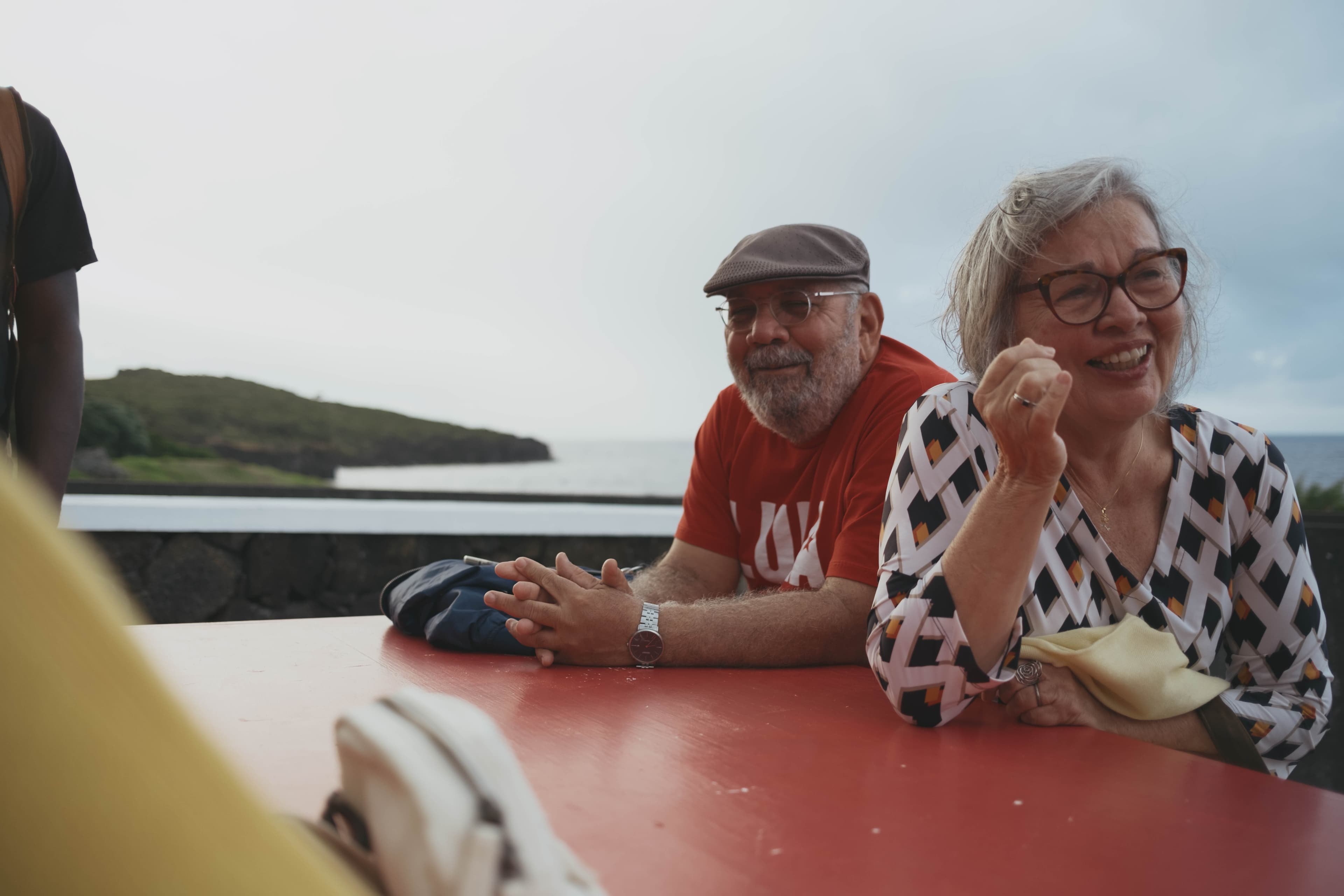 Couple sitting at red outdoor table on boat deck in the Azores. Man wears brown flat cap, glasses, red hoodie, and watch, with hands clasped on table. Woman wears glasses and geometric patterned top, touching her face while smiling. Green hills and coastline visible across calm water in background under overcast sky.