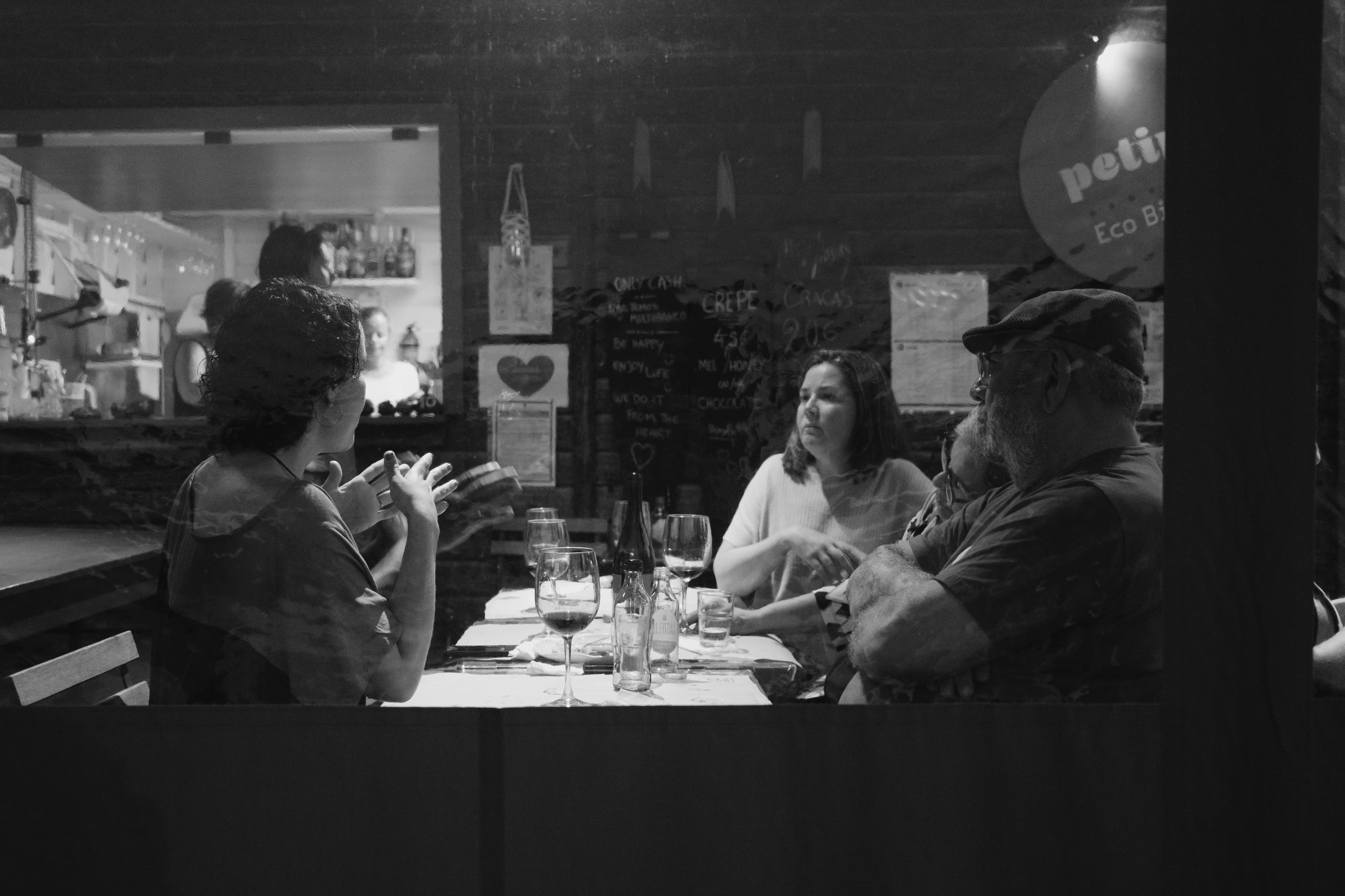 Black and white photo of three people dining at rustic restaurant table in the Azores. Woman on left gestures while speaking, woman in center wears white sweater, bearded man on right wears knit cap. Table set with wine glasses, water glasses, and napkins. Stone walls decorated with chalkboard menu, framed photos, and hanging items create cozy restaurant atmosphere.