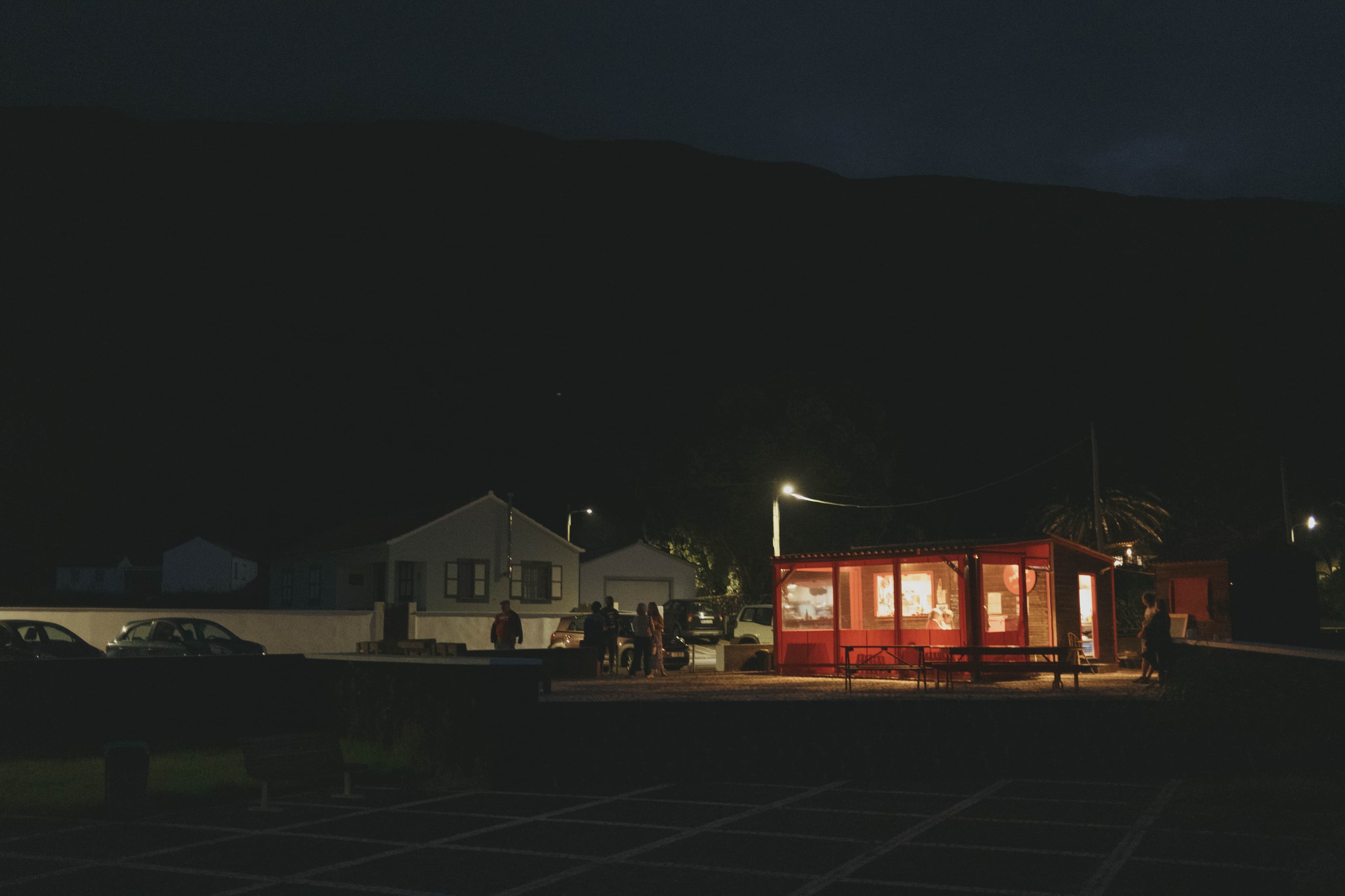 Night scene in Azores village showing brightly lit red restaurant or shop with large windows glowing warmly against dark sky. White residential houses visible in background, with parked cars and small groups of people gathered outside the illuminated building. Street lights create pools of light in the darkness, palm tree silhouette visible on right side.
