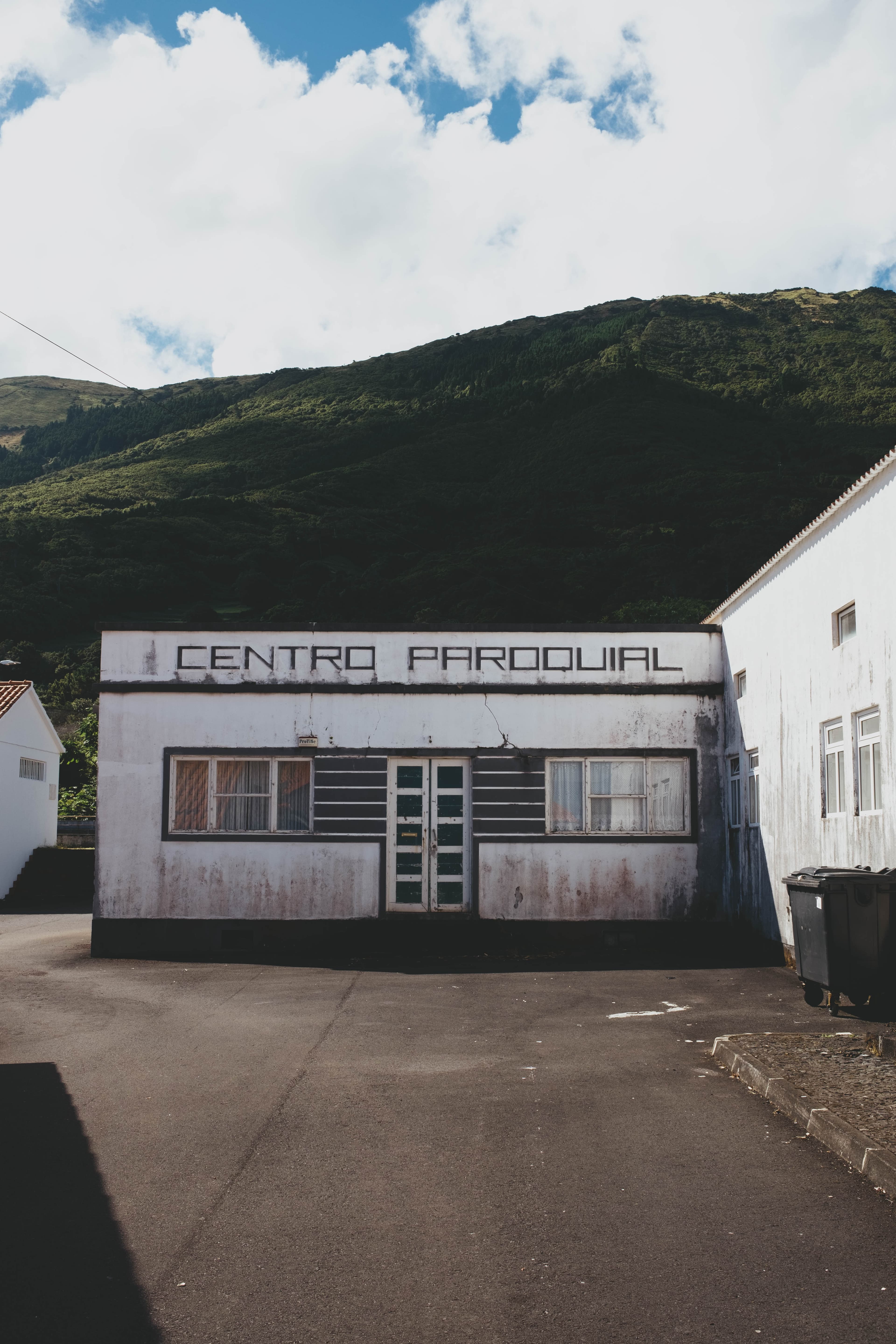 Weathered white building labeled 'CENTRO PAROQUIAL' (Parish Center) in an Azorean village, nestled against lush green volcanic hills. The single-story structure features wooden shuttered windows, a central glass door, and shows signs of age with stained walls. Traditional Portuguese tile roofing and adjacent white buildings complete the rural community setting under a partly cloudy sky. The parish center sits on a concrete plaza, serving as a focal point for local community gatherings.