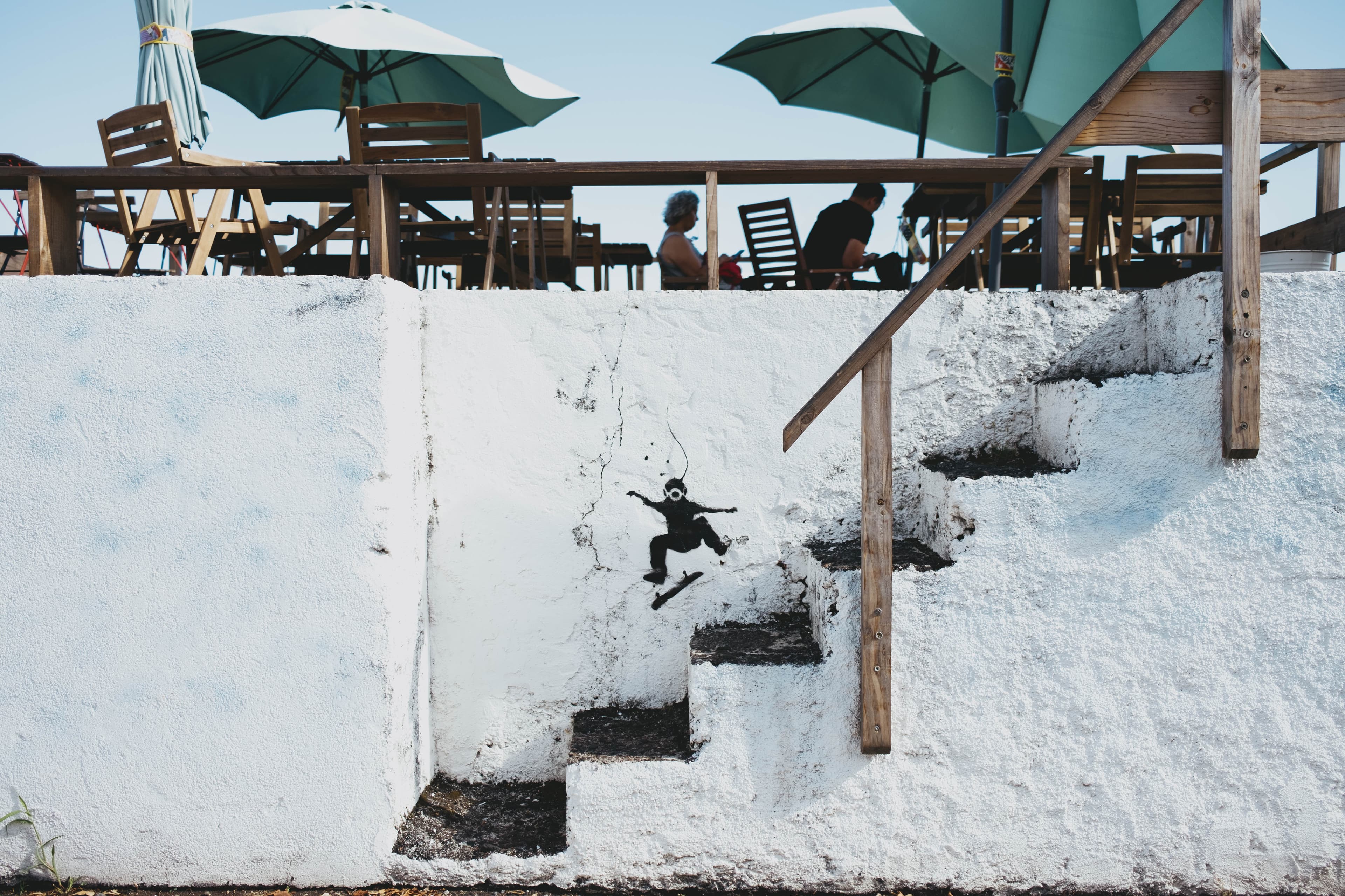 Elevated wooden deck restaurant terrace in the Azores built above white-painted volcanic rock formations. Green and white umbrellas shade wooden tables and chairs where diners are seated. A wooden staircase descends along the cliff face, and black stencil street art of a skateboarding figure decorates the white-washed rock wall. The unique dining setup utilizes the natural volcanic coastline architecture typical of Azorean seaside establishments.