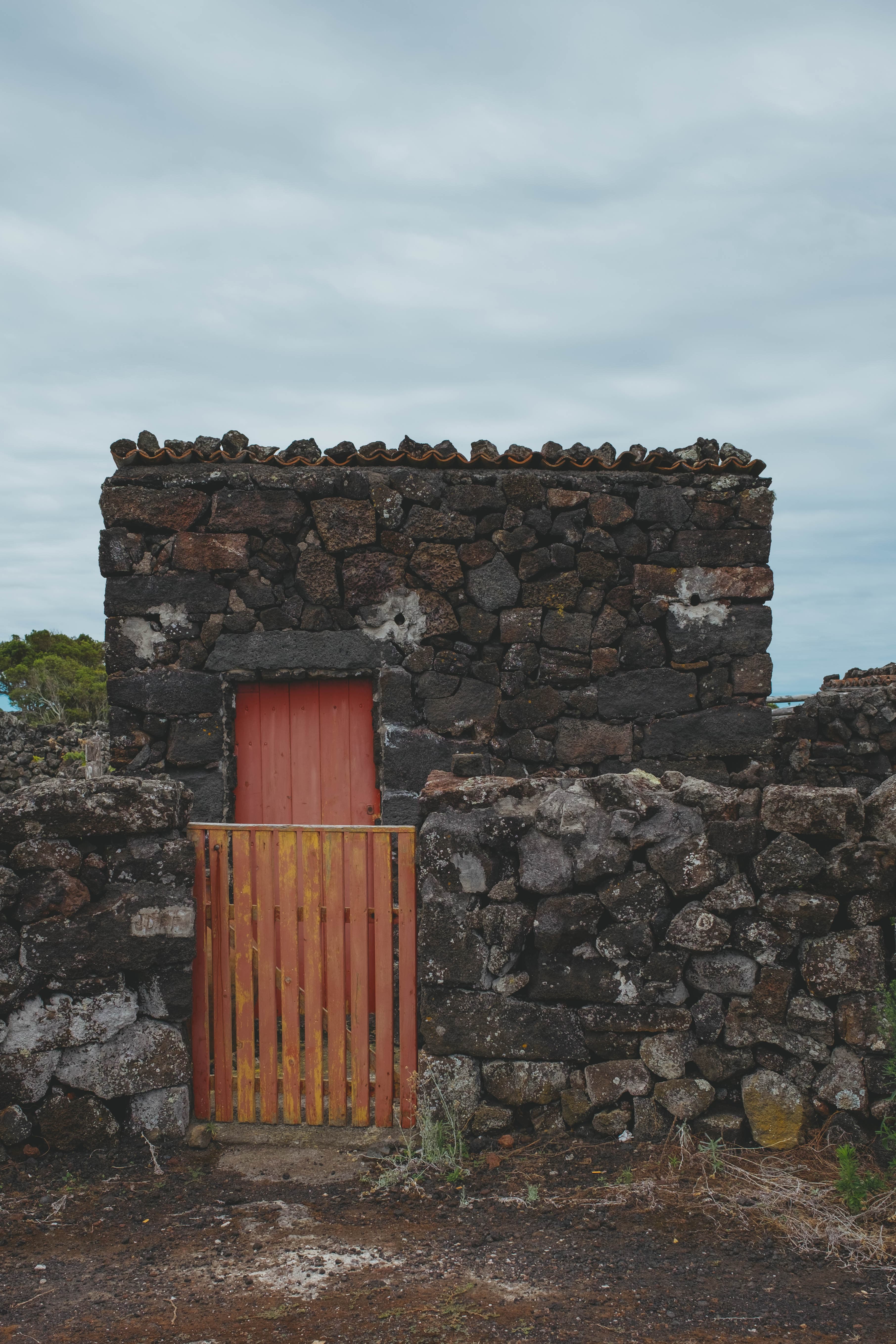 Traditional Azorean stone house with volcanic rock walls and terracotta tile roof, featuring a weathered wooden gate and door in rustic orange-red tones against an overcast sky on Pico Island.