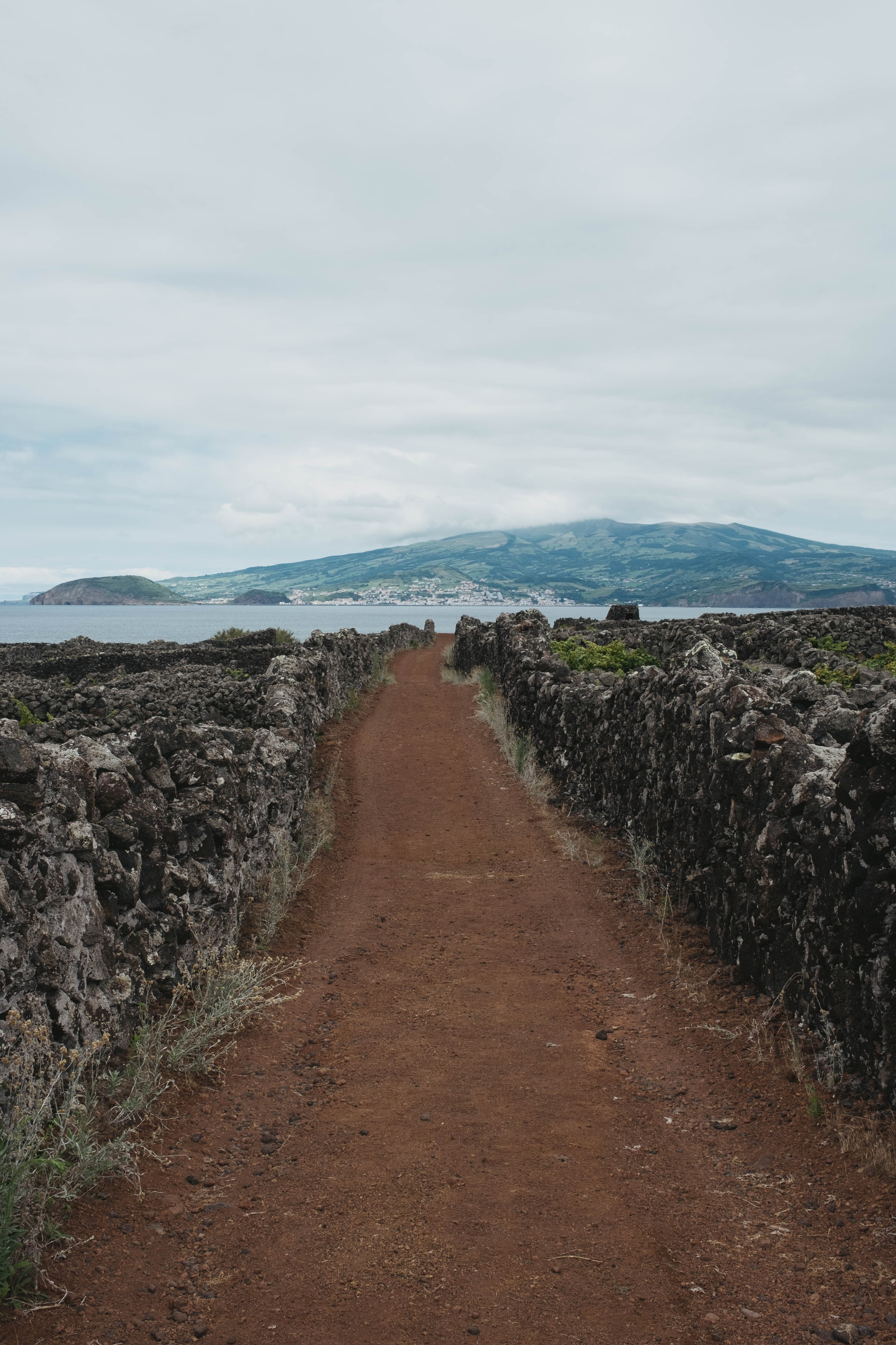 Red dirt path flanked by traditional volcanic stone walls leading toward the ocean, with Faial Island visible across the channel under cloudy skies.
