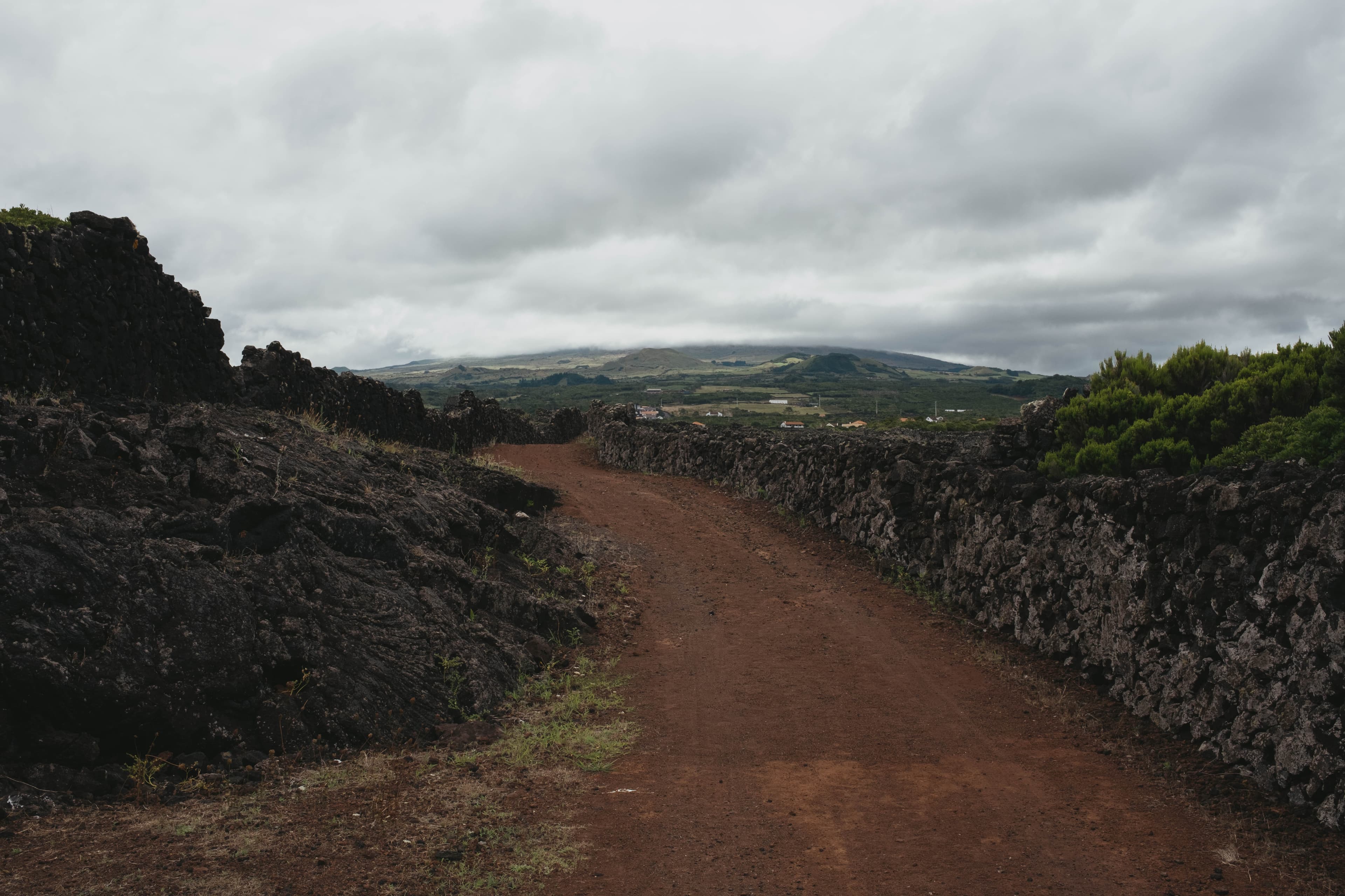 Dirt path winding through dramatic volcanic rock formations with Pico Mountain's slopes visible in the distance under moody overcast skies.