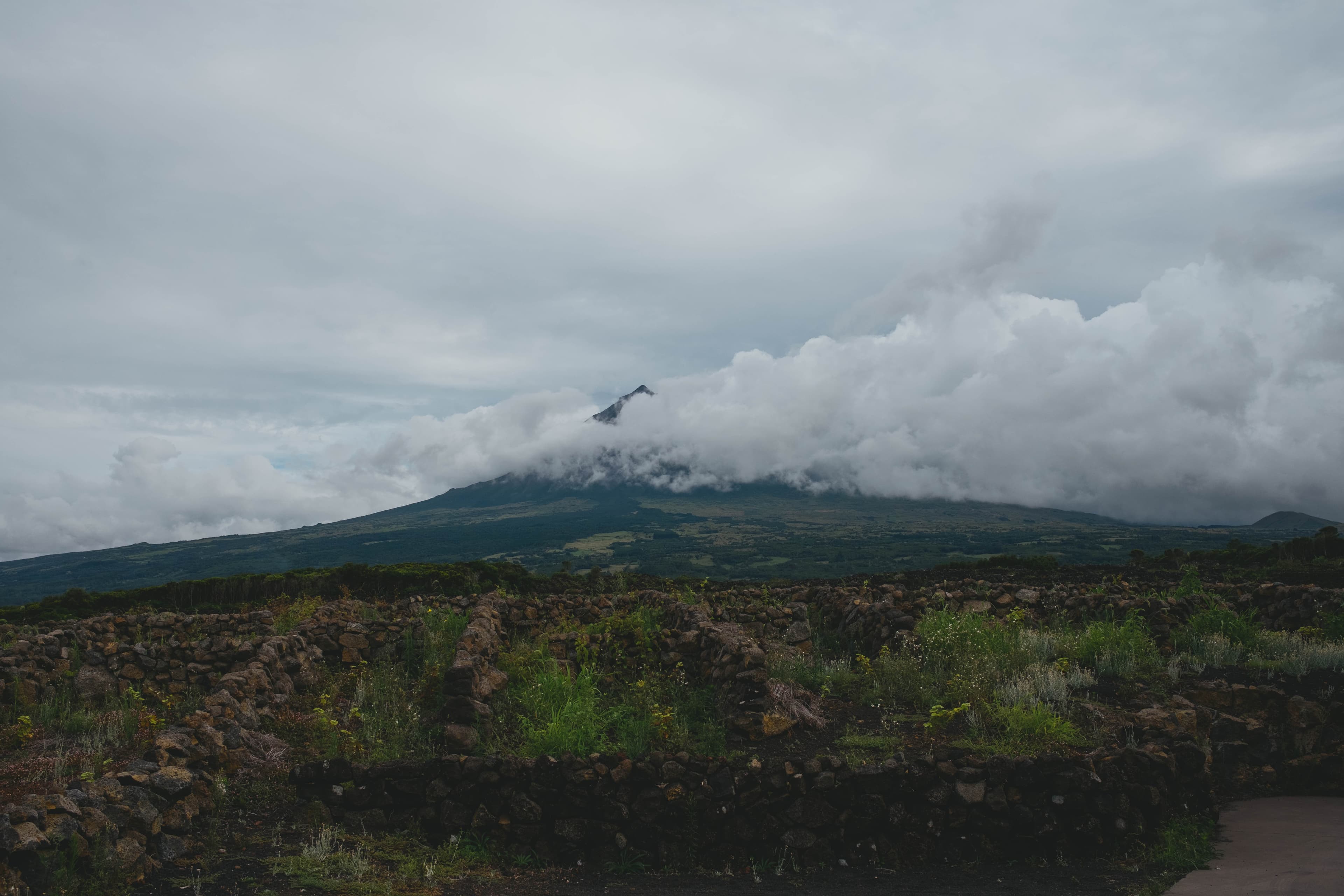 Mount Pico shrouded in dramatic clouds above a rocky lava field dotted with green vegetation under an overcast sky