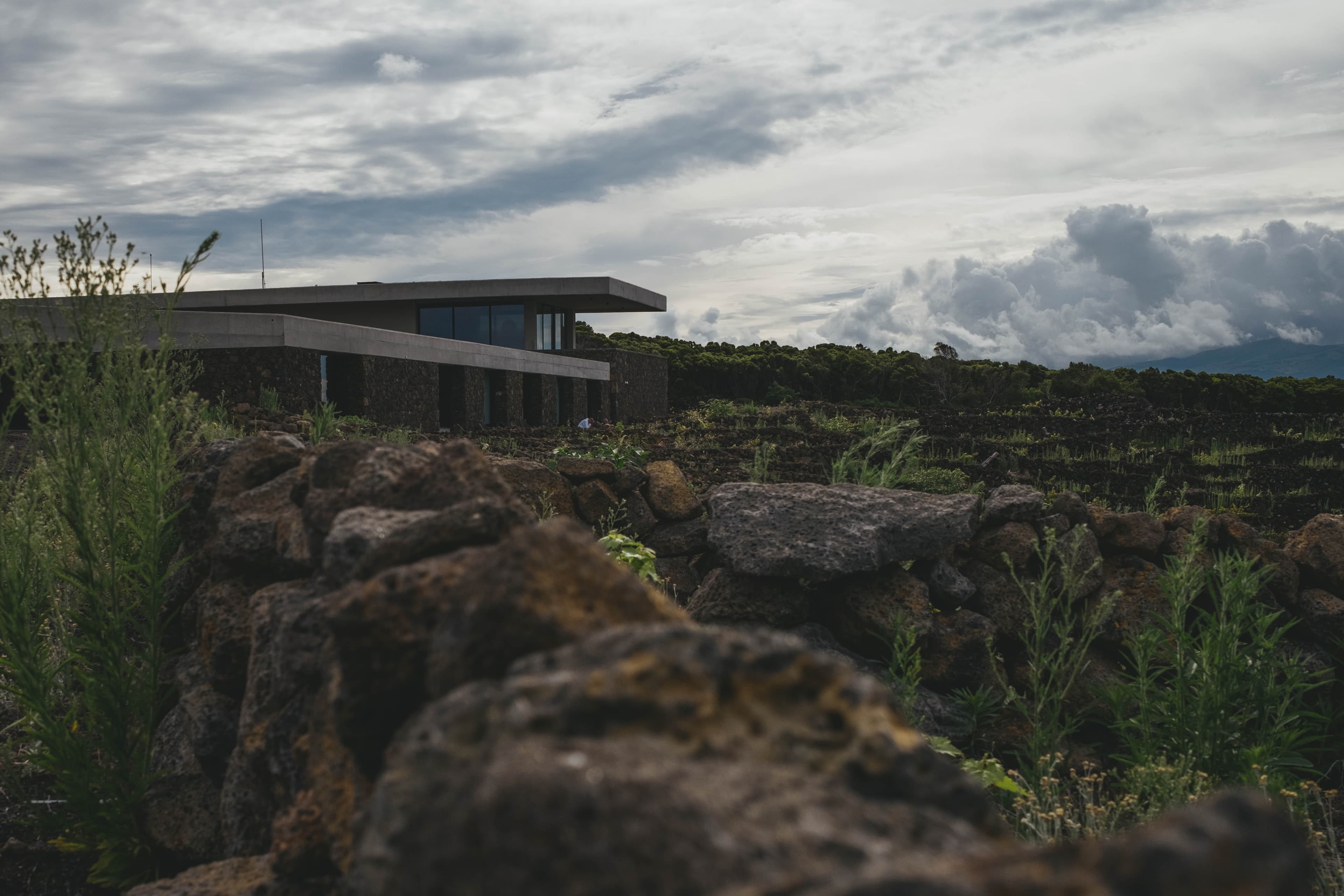 Modern concrete winery building of Azores Wine Company set among volcanic rock formations and traditional stone-walled vineyard plots under dramatic cloudy skies